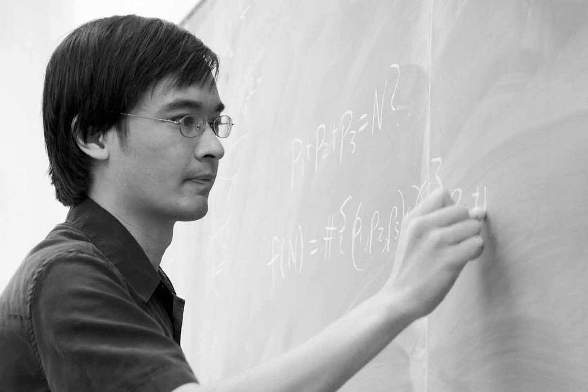  Black-and-white photo of a person writing mathematical formulas on a chalkboard.