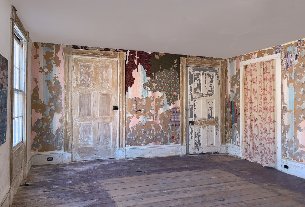 Installation view showing layered mixed textiles integrated with existing wallpaper in a weathered interior room.