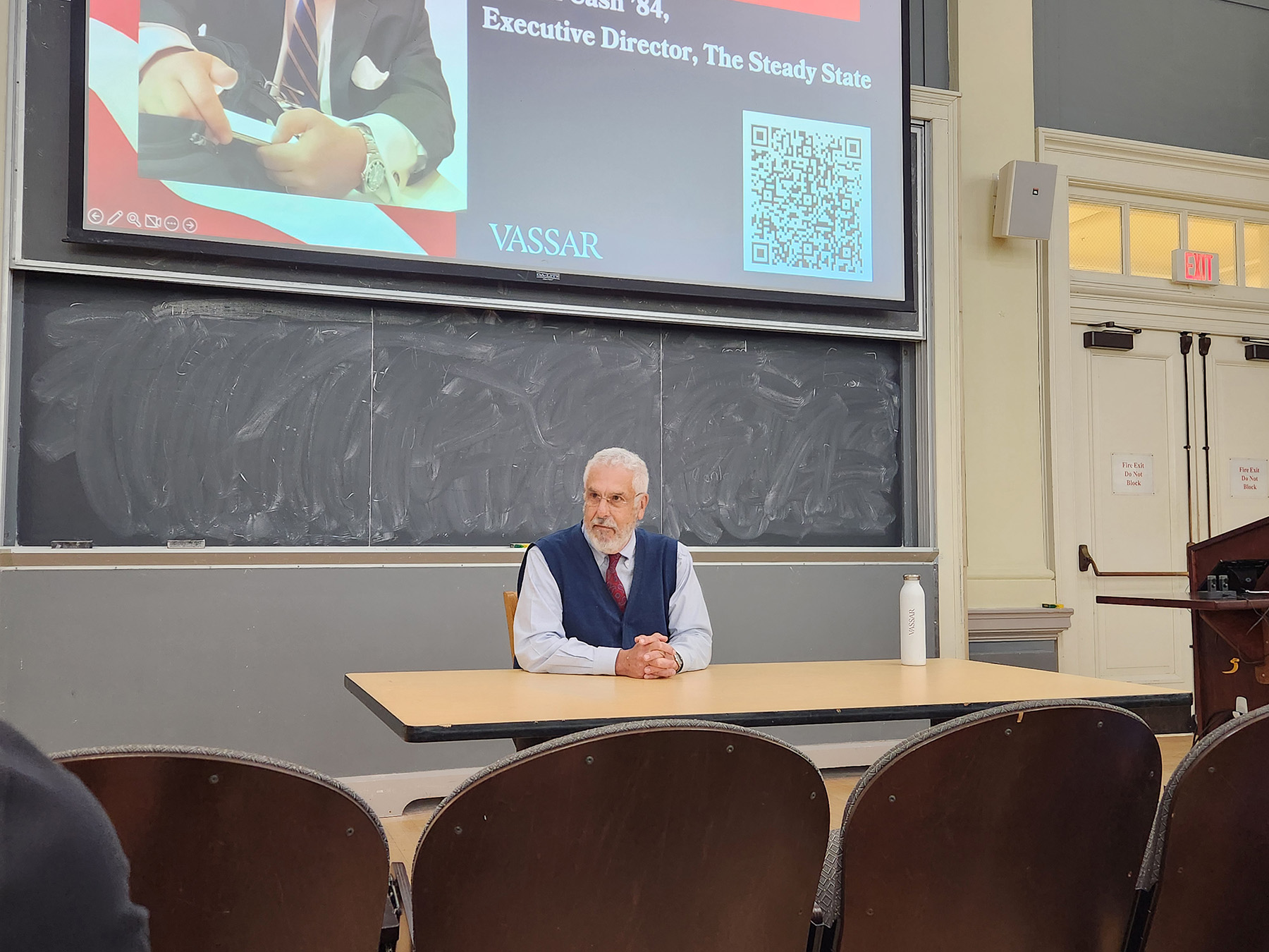 Person seated at a classroom table in front of a chalkboard, with a Vassar presentation slide and QR code projected above.