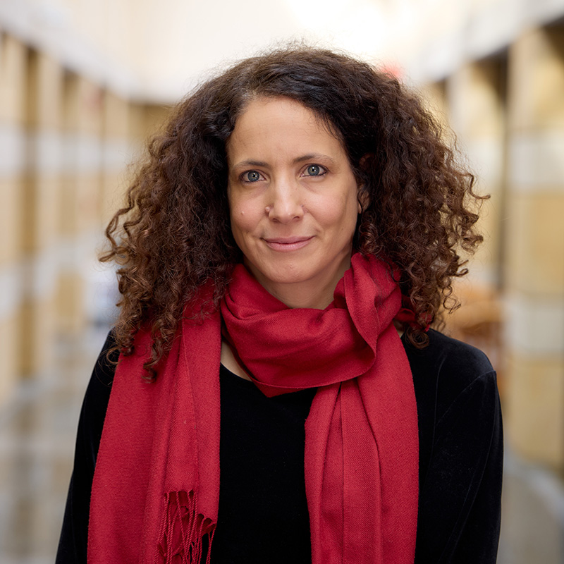 A person with long, curly hair stands in a hallway, wearing a black top and a red scarf, looking directly at the camera.