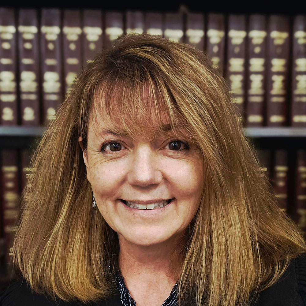  Hon. Joan M. McMenemy with shoulder-length light brown hair, wearing a dark top, in front of shelves filled with large books.