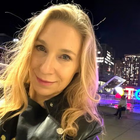 Sheena Josselyn with long blonde hair wearing a black jacket, smiling at night with city lights and illuminated buildings in the background.