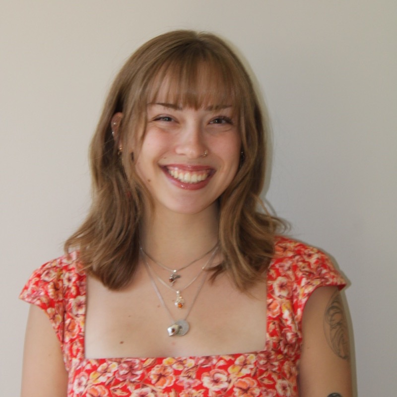 A person with shoulder-length wavy hair and bangs smiles in front of a plain, light-colored wall. They wear a red floral-patterned top with a square neckline and layered necklaces. A tattoo is partially visible on one upper arm. The lighting is soft and evenly distributed.