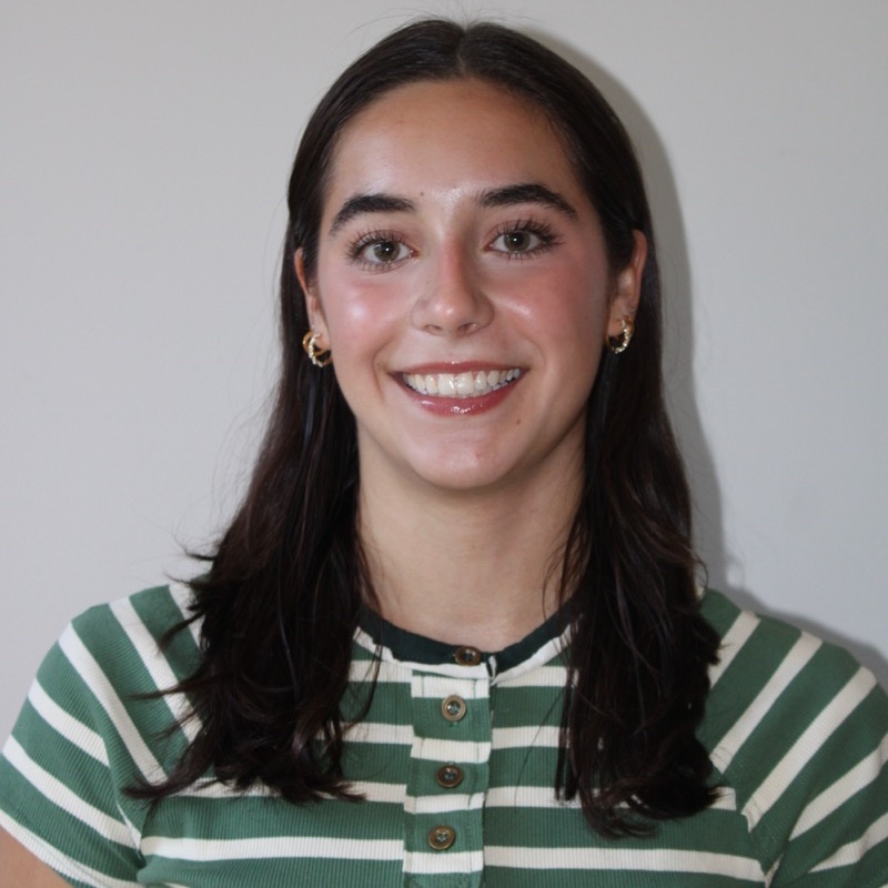 A person with straight, dark hair parted in the middle smiles while standing against a neutral background. They wear a green and white striped button-up shirt and small hoop earrings. Their posture is relaxed and centered. Lighting highlights the face with soft shadows.
