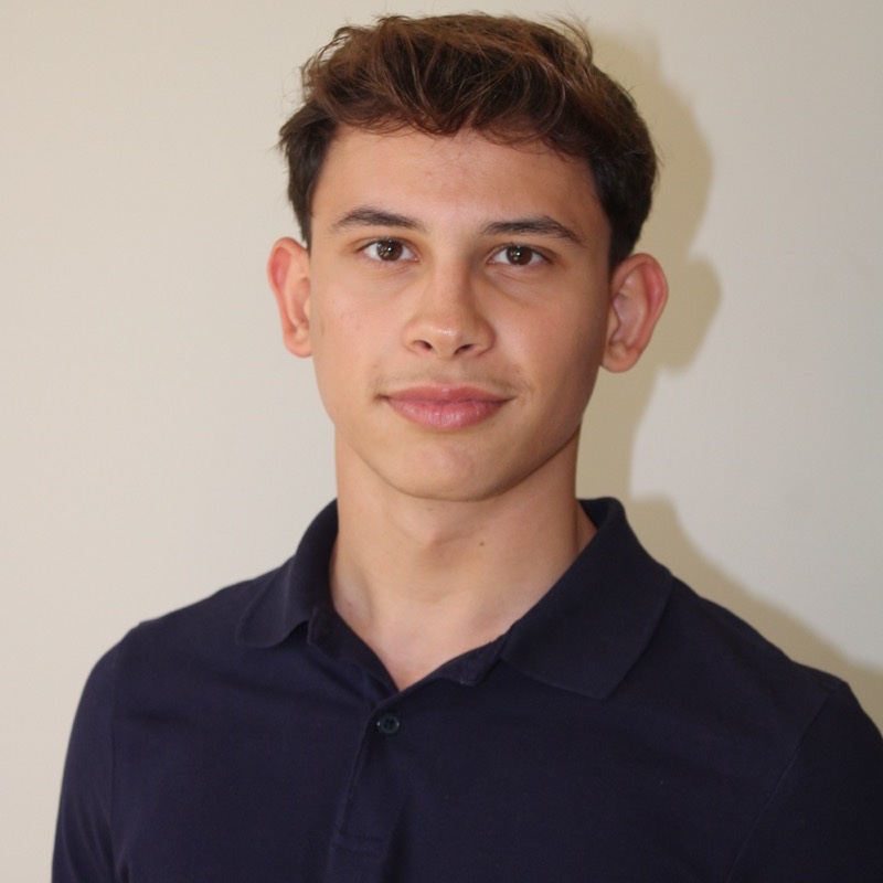 A person with short, dark, tousled hair stands in front of a light-colored wall. They wear a navy blue collared shirt and look directly at the camera with a neutral expression. The image is well-lit, with light coming from the front and slightly to the side. Shadows are minimal.