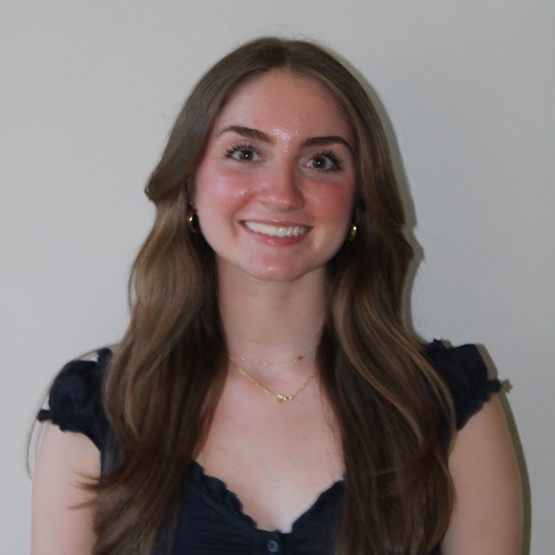 A person with long, loosely curled hair smiles in front of a plain backdrop. They wear a navy blue top with ruffled sleeves and delicate jewelry including a short necklace and small hoop earrings. The lighting is soft and even. Their body is angled slightly while facing the camera.