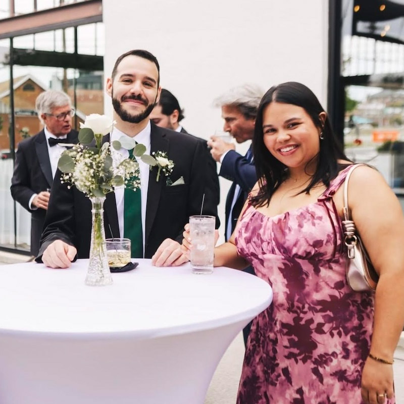  Two individuals smile while standing at a cocktail table. The person on the left wears a black suit with a green tie and a white boutonnière. The person on the right wears a pink patterned dress and holds a drink. Others are visible in the background.