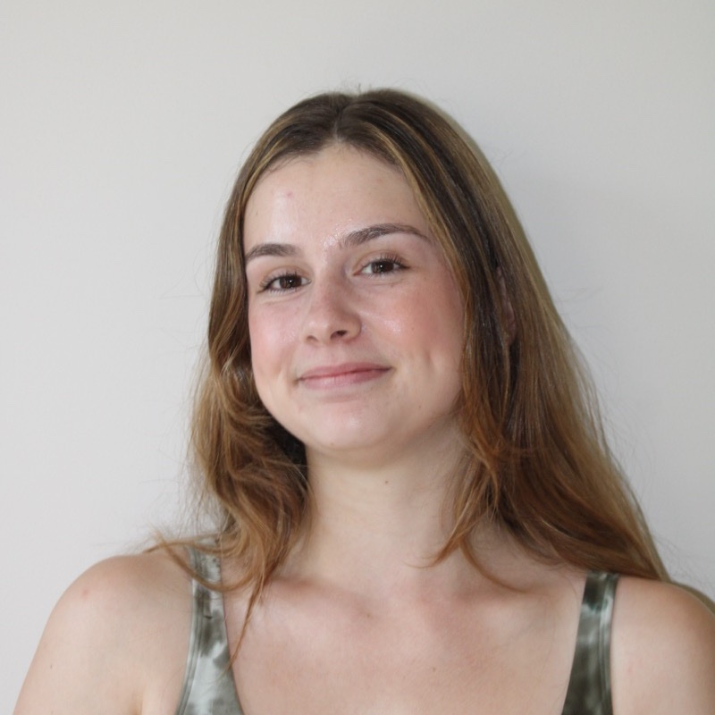 A person with long, light brown hair poses in front of a pale wall. They wear a green tie-dye tank top and have a subtle smile. The lighting is natural and even, with slight shadowing along one side. The composition is centered and minimal.