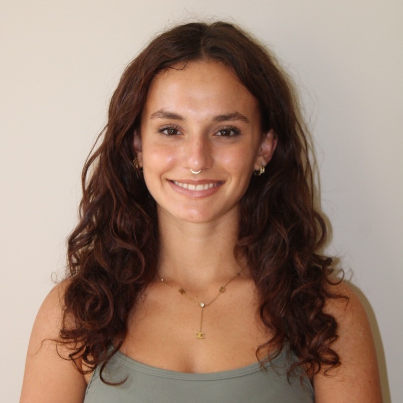 A person with long, curly hair stands against a neutral wall, smiling at the camera. They wear a sage green tank top, gold hoop earrings, and a pendant necklace. A septum ring is visible. Lighting is soft and evenly fills the frame.