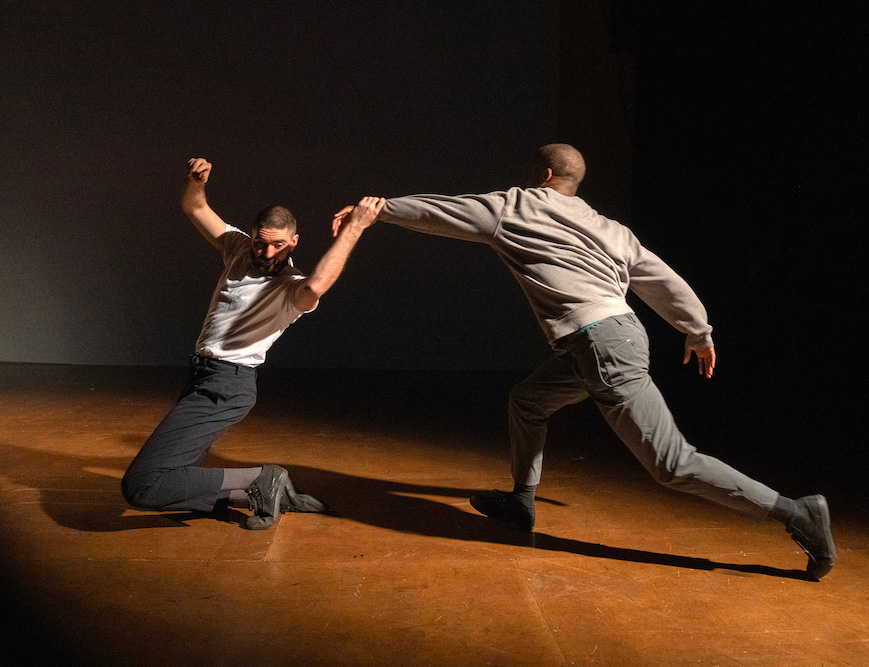 Two dancers perform on stage; one kneels with an arm raised while being pulled forward by the other, who leans away with an outstretched arm.