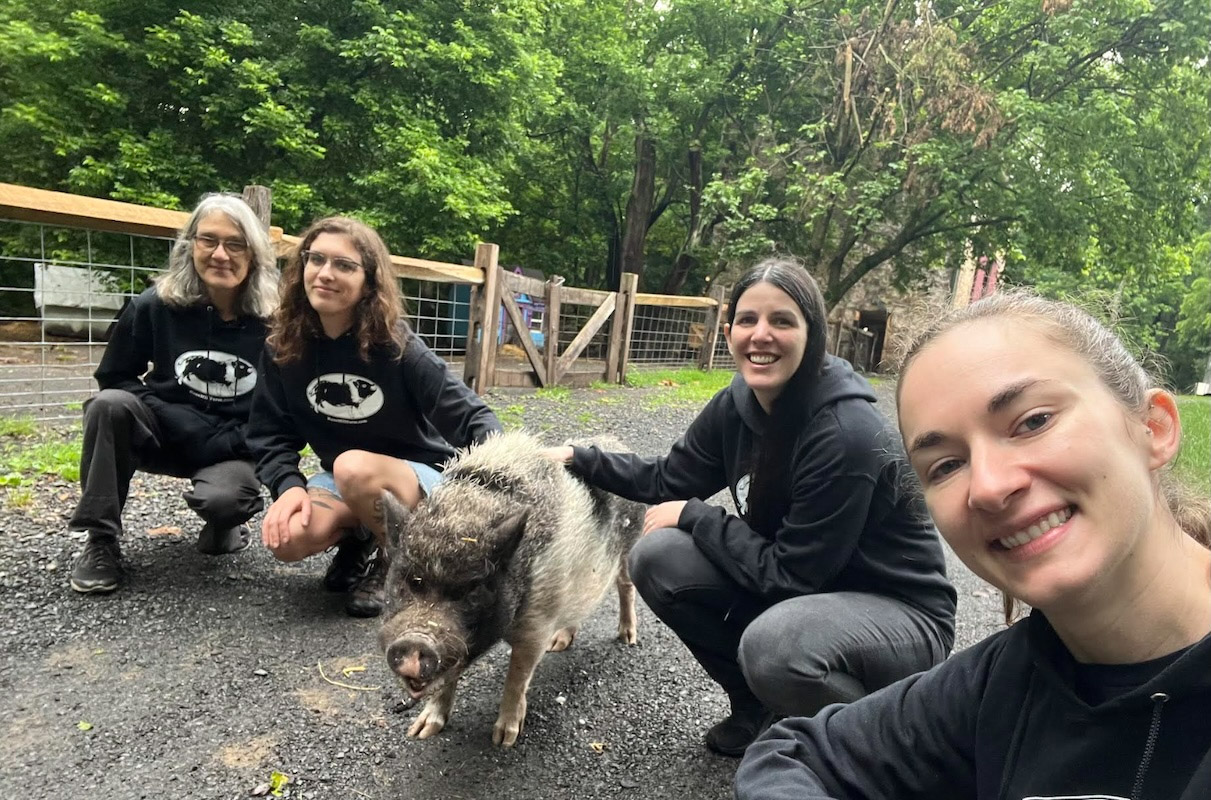  Four individuals crouch on a dirt path with a hairy pig. The pig is the focus, with three people smiling and looking toward the camera. They are all wearing matching black sweatshirts with a pig logo on them. Trees and a wooden fence are in the background.
