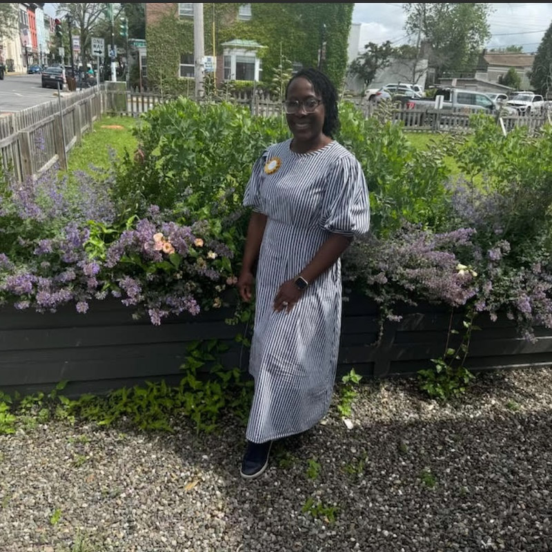  A person stands in front of a raised garden bed filled with green plants and purple flowers. They are wearing a striped dress with puffy sleeves and an orange pin on their shoulder. A street with a building and cars is visible in the background.