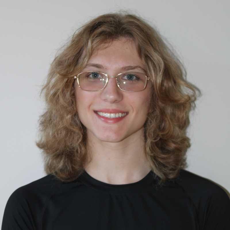 A person with shoulder-length curly hair and glasses smiles in front of a light background. They wear a black shirt and gold-rimmed eyeglasses. Lighting is soft, highlighting facial features evenly with minimal shadow. The composition is symmetrical and centered.