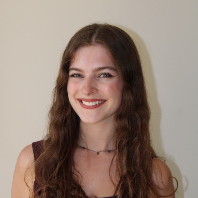 A person with long, wavy brown hair smiles slightly to the side. They wear a sleeveless burgundy top and a simple black necklace. The light is warm and directional, casting a soft shadow on the wall behind. The overall tone is casual and clear.