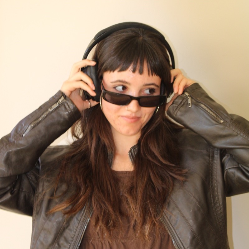 A person wears a dark leather jacket and large over-ear headphones while adjusting them with both hands. They also wear dark sunglasses and a brown top, with long dark hair and short bangs. The background is plain, and lighting is warm with soft highlights. Their gaze is turned slightly to the side.