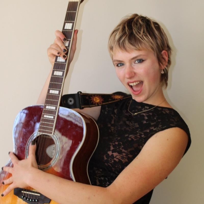 A person with short, tousled hair holds an acoustic guitar and poses with an open-mouthed expression. They wear a black lace top and a strap with sun designs is visible over one shoulder. The guitar is angled forward and brightly polished. Lighting is direct, creating high contrast and sharp details.
