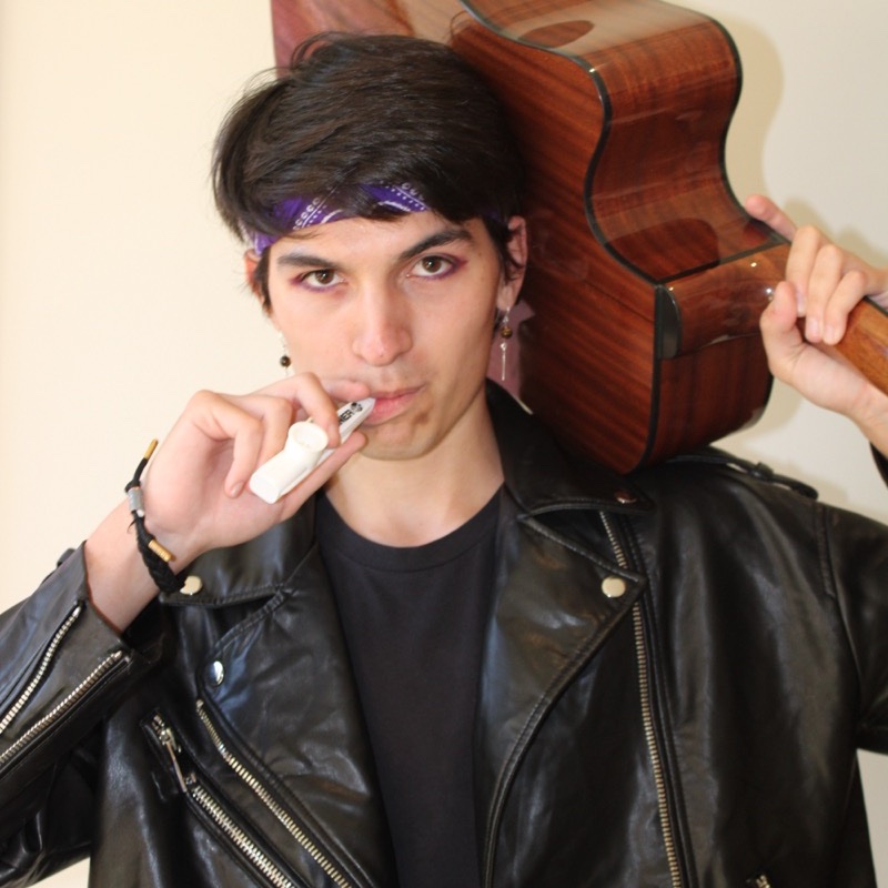 A person in a black leather jacket holds an acoustic guitar over one shoulder and a vape pen to their mouth. They wear a purple bandana tied around their head and a black shirt underneath the jacket. Their gaze is direct, and their expression is neutral. The background is softly lit and neutral in color.