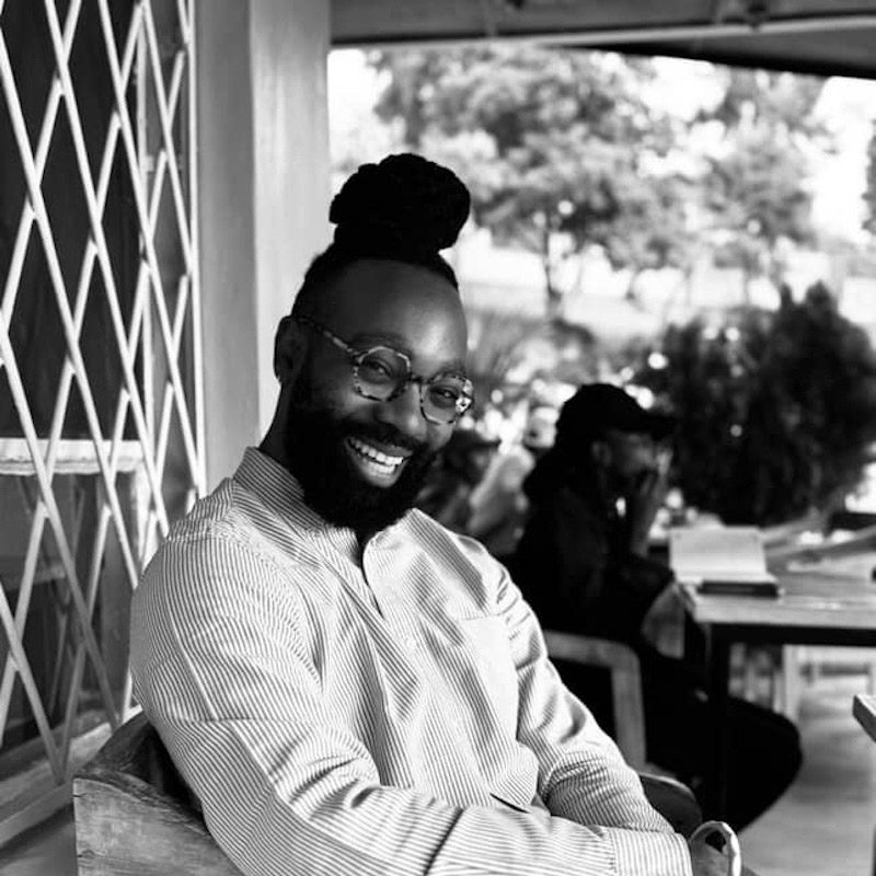  A person with a beard and hair in a bun smiles at the camera while sitting in a chair. The person wears round glasses and a striped shirt. This black-and-white photo is taken in a casual, outdoor setting with another person visible in the background.