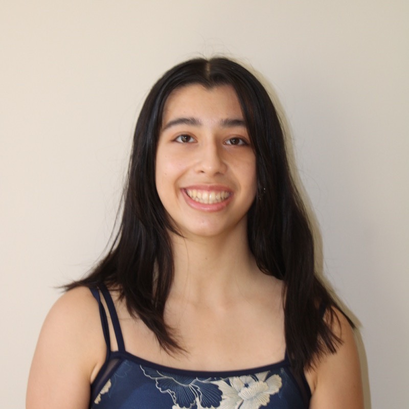 A person with long dark hair stands against a light-colored wall, smiling with their teeth showing. They wear a navy blue camisole top with a white and beige floral pattern. Their posture is upright and relaxed. The lighting is even and soft, with minimal shadow.