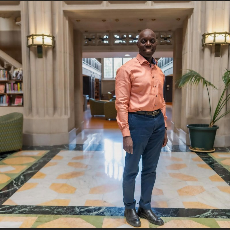  A person stands in the center of a well-lit, ornate hallway with a patterned tile floor. They are wearing an orange shirt, dark pants, and black shoes. The background includes vaulted ceilings, wooden archways, and a glimpse of a library.