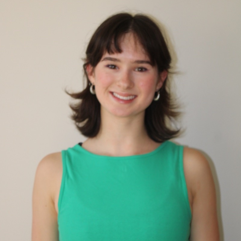 A person with short, softly curled dark hair smiles while standing in front of a pale wall. They wear a sleeveless green top and silver hoop earrings. Their expression is gentle, and their posture is relaxed. The image is softly lit with natural tones.