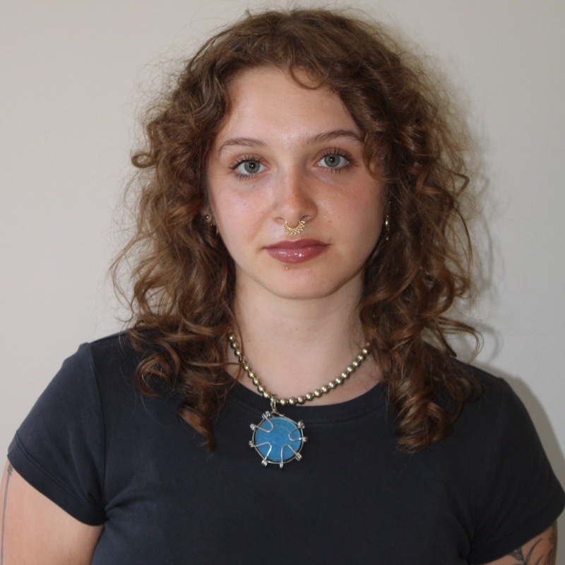 A person with shoulder-length curly hair wears a dark t-shirt and a large beaded necklace with a bright blue pendant. They have a septum ring and a neutral facial expression while facing the camera directly. The lighting is even, showing texture and detail in both clothing and hair. The background is plain and softly lit.
