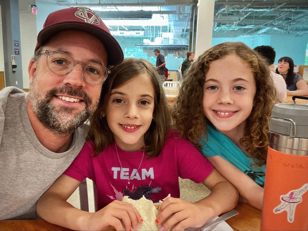  A person wearing a baseball cap and glasses takes a selfie with two children. All three are smiling and sitting at a table with food. The background is a large, open indoor space with other people.