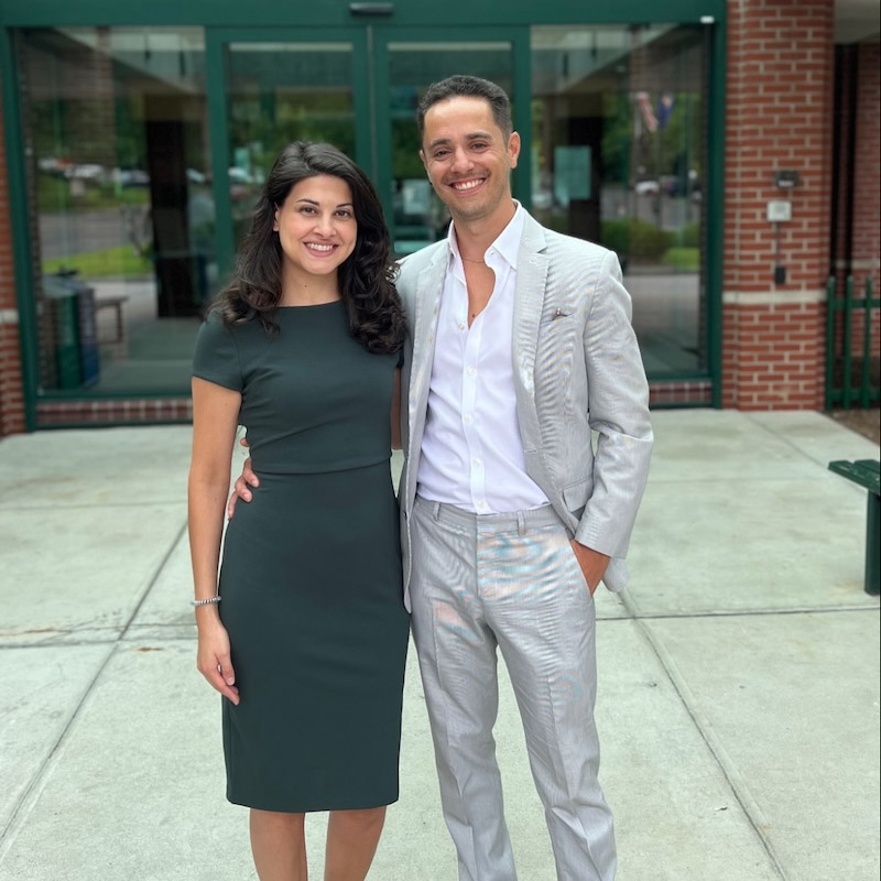 Two individuals stand side-by-side, smiling for the camera. The person on the left wears a dark green dress, and the one on the right wears a light gray suit with a white shirt. They are outdoors on a paved area in front of a brick building with glass doors.