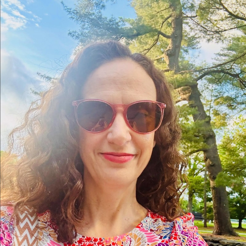  A person with wavy brown hair smiles at the camera while wearing pink sunglasses and a brightly colored floral top. The person is outdoors, with tall trees and a sunny sky in the background.