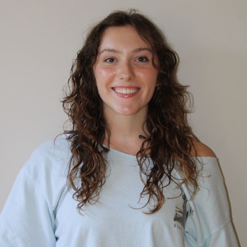 A person with long curly hair smiles in front of a light-colored wall. They wear an oversized light blue t-shirt with one shoulder exposed. The lighting is natural, creating soft shadows and highlights. Their posture is relaxed and centered.