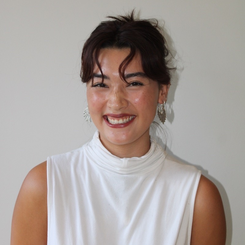 A person with dark hair styled in a loose updo smiles broadly at the camera. They wear a sleeveless white top with a high neckline and large, detailed earrings. The background is light and evenly lit. Their expression is joyful and the composition is centered.