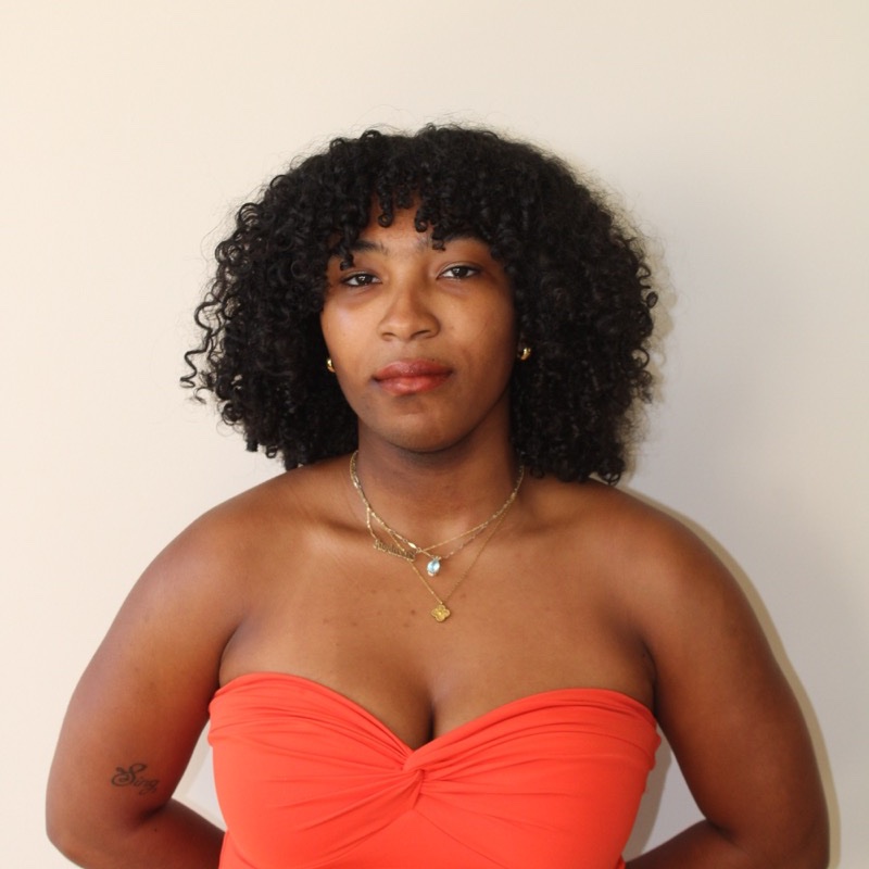 A person with medium-length curly hair stands in front of a pale wall, wearing a strapless orange-red top. They have a neutral expression and wear layered gold necklaces and small hoop earrings. A small tattoo is visible on one upper arm. The lighting is warm and natural.
