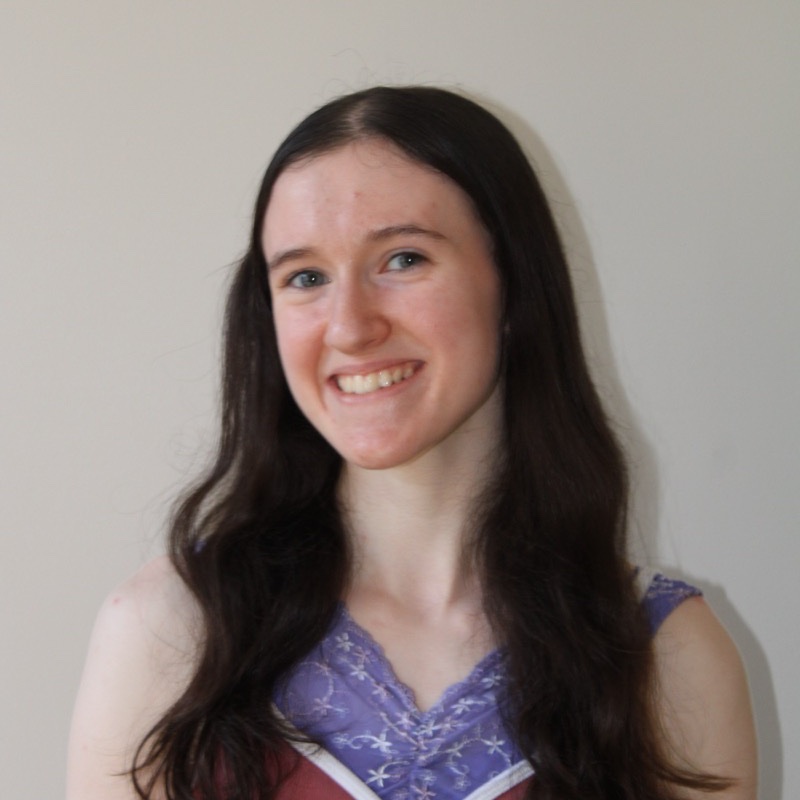 A person with long dark hair smiles while standing in front of a plain background. They wear a purple and maroon sleeveless top with decorative stitching along the neckline. Their posture is upright and slightly angled. Lighting is soft and natural.