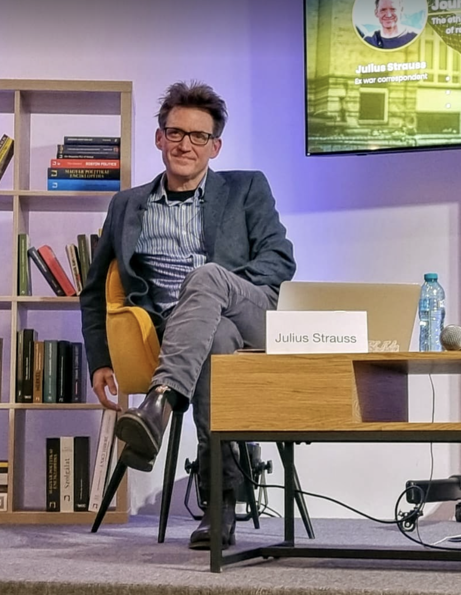 Julius Strauss, a person with short, tousled dark hair and glasses, is seated in a mustard-yellow chair with one leg crossed over the other. They wear a dark blue blazer, a striped shirt, grey pants, and black boots. To their right is a wooden table with a name card that reads "Julius Strauss," a plastic water bottle, and a microphone. Behind them is a bookcase filled with colorful books, and a screen displaying their photo, name, and the title "Ex war correspondent."