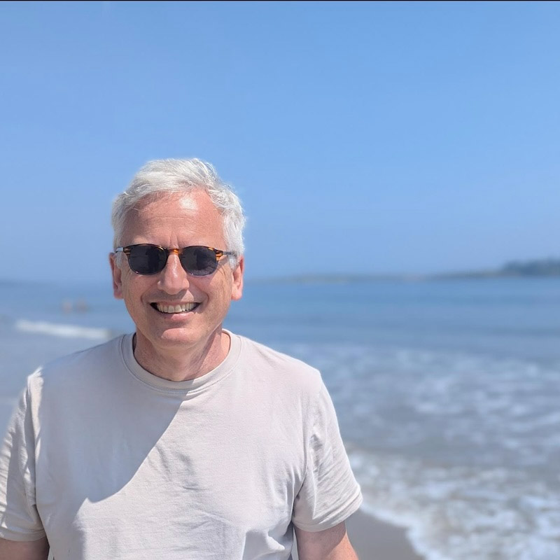 A person with short, light-colored hair and tortoiseshell sunglasses stands on a sandy beach, smiling at the camera. The ocean is behind them with a shoreline visible in the distance. The sky is clear and blue.
