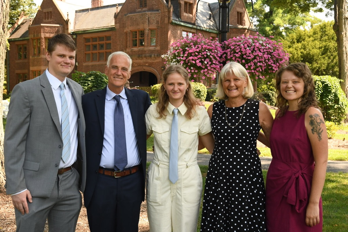 A group of five individuals stands outside in front of a brick building with blooming flowers. From left to right: one person in a gray suit, two in dark blue suits, and two in dresses—one black with white polka dots and one a deep red.