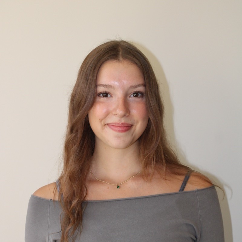 A person with long wavy hair stands in front of a neutral wall, smiling softly. They wear a gray off-the-shoulder top and a delicate necklace with a small green pendant. The lighting is even, and the tone of the image is calm and minimal. Their posture is centered and relaxed.