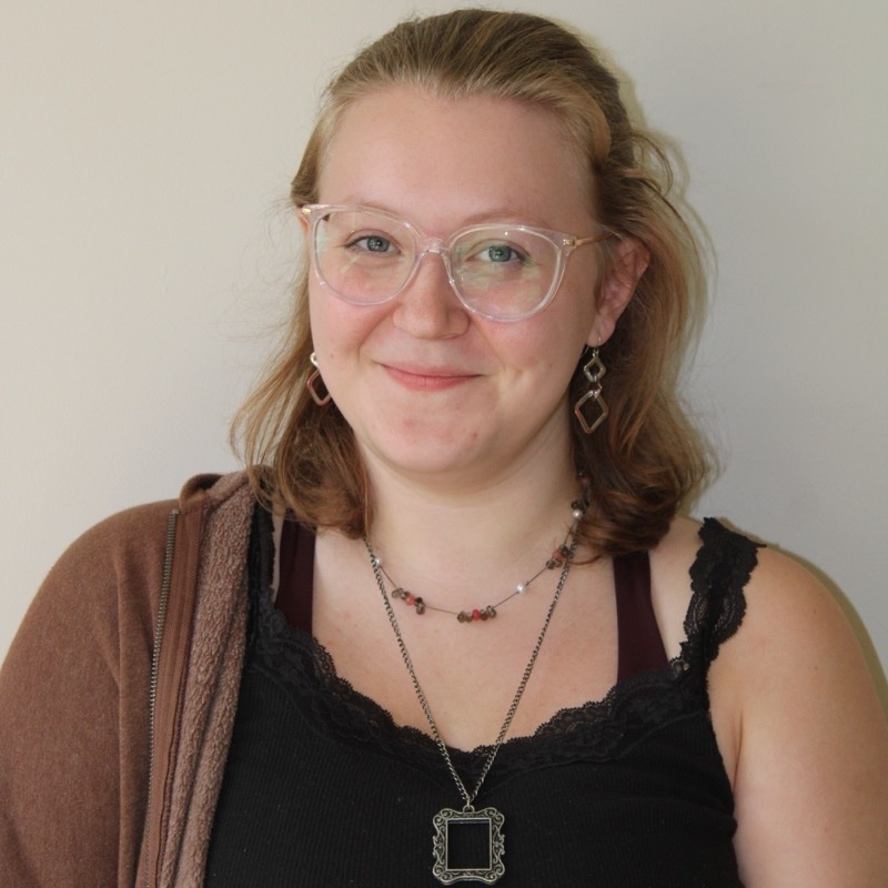 A person with shoulder-length hair wears translucent glasses, geometric earrings, and layered necklaces, including one with an ornate pendant. They wear a black lace-trimmed top and a brown zip-up sweater off one shoulder. Their expression is soft and smiling. The lighting is even, with a plain wall in the background.