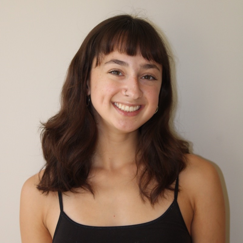 A person with medium-length dark brown hair styled in loose waves smiles in front of a light wall. They wear a thin-strapped black top and small hoop earrings, with a nose ring visible on one side. Their expression is friendly, and the lighting is soft and natural. The framing is centered and clear.