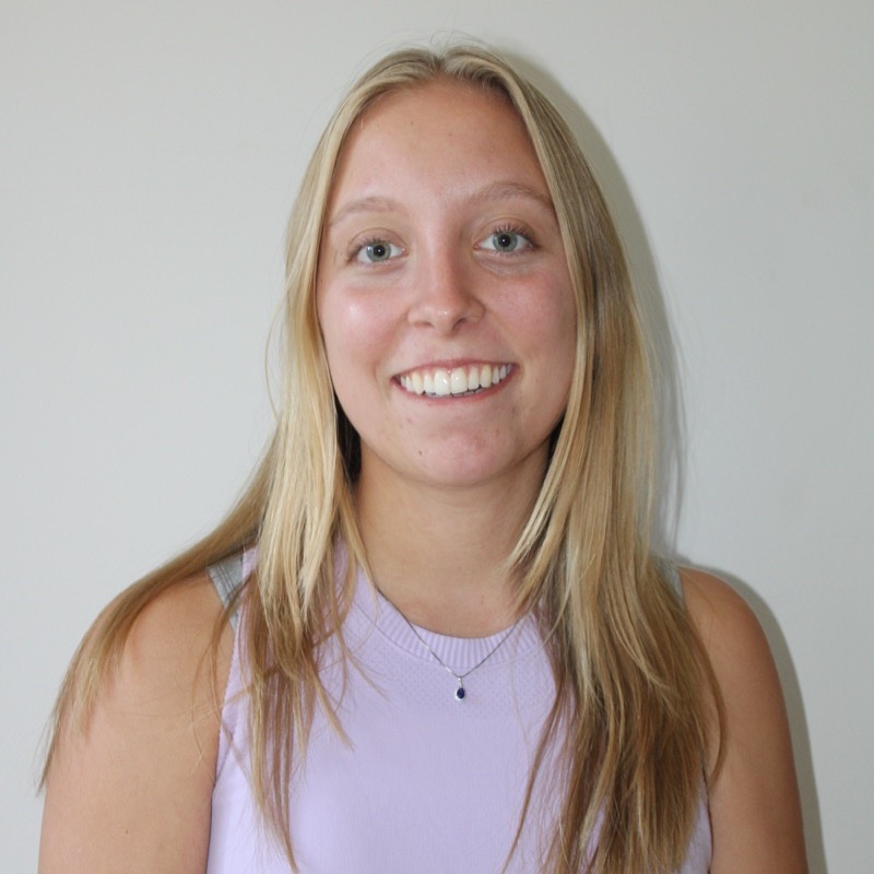 A person with long, straight blonde hair smiles broadly in front of a plain, light-colored wall. They wear a lavender sleeveless top and a small necklace with a dark pendant. The lighting is bright and even, casting minimal shadows. Their posture is upright and centered.