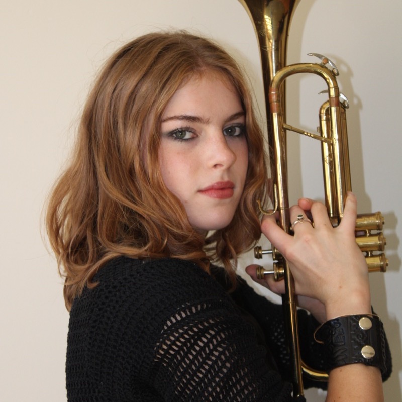 A person with shoulder-length wavy hair holds a brass trumpet upright next to their face. They wear a black, open-knit top and a thick black wristband with metal details. Their expression is serious and direct. The lighting is bright and highlights the shiny instrument.