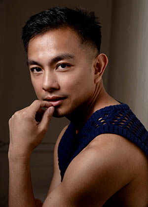A portrait of Norbert de la Cruz III. They are seated in a three-quarter pose, turned slightly to the side with one hand resting against their chin. They have short dark hair and wear a sleeveless, dark blue knitted top. The lighting is soft and directional, casting gentle shadows that define the contours of the face and shoulder. The background is plain and dimly lit, keeping the focus on the subject.