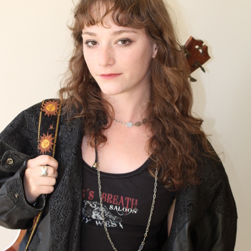A person with long, curly hair wears a black jacket with a sun-patterned guitar strap over one shoulder. They have a calm expression and wear layered necklaces, including a chain with round discs and another with a pendant. A guitar headstock is partially visible behind them. The lighting is soft and warm.
