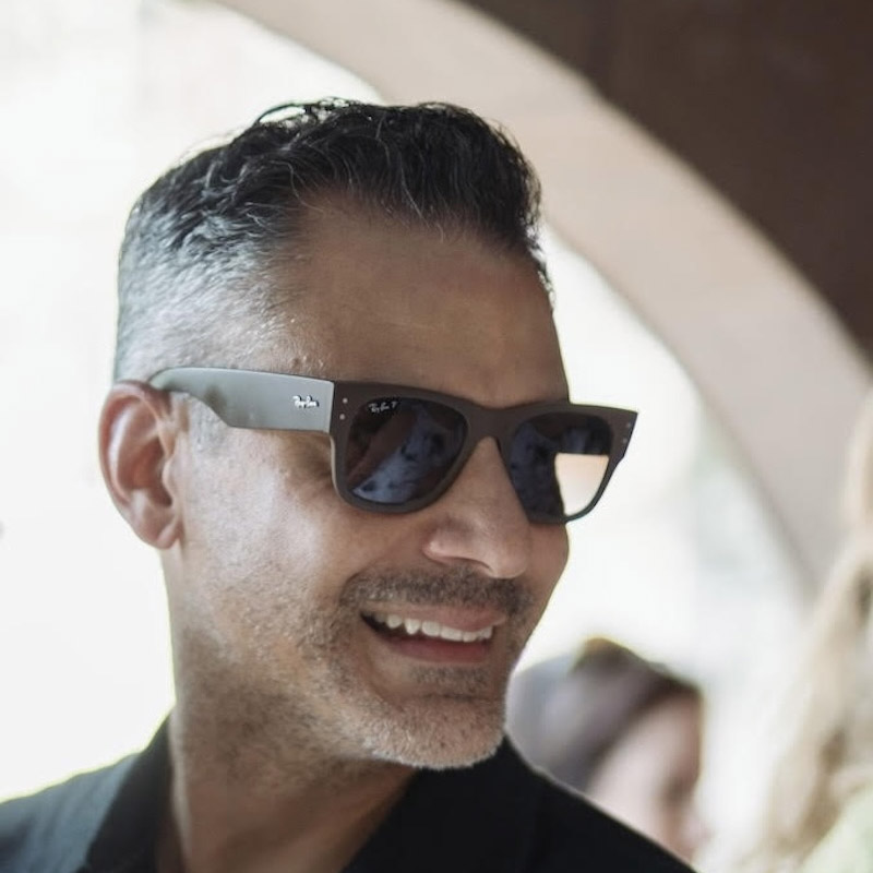  A person with a styled haircut and sunglasses is smiling. The person is looking to the side, away from the camera. The background is slightly out of focus, suggesting an indoor or shaded outdoor setting.
