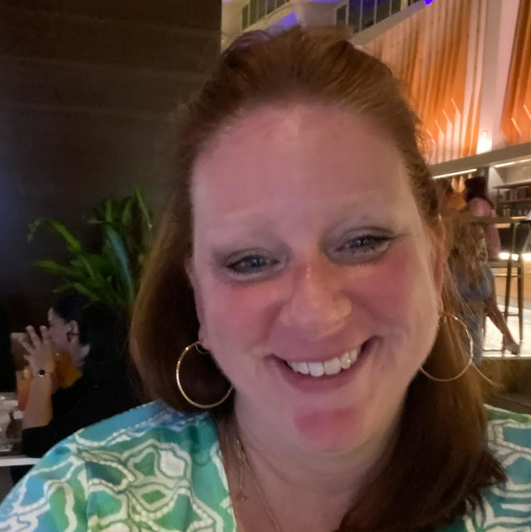  A person with reddish hair and large hoop earrings smiles directly at the camera. The background is a dimly lit indoor setting with other patrons and modern decor.