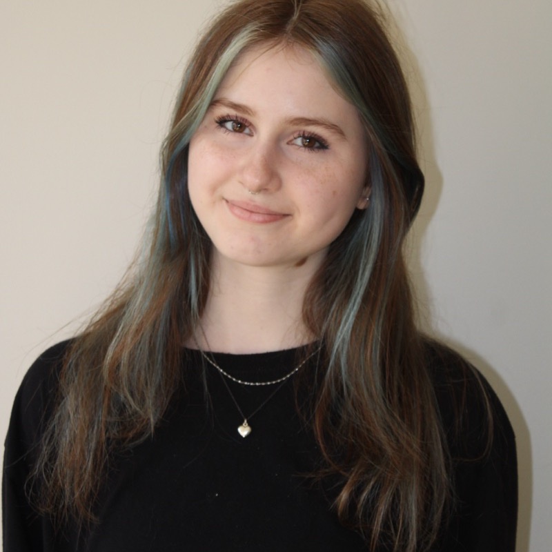 A person with long hair dyed with light blue streaks wears a black top and layered necklaces, one of which has a small heart-shaped pendant. Their expression is neutral with a slight smile. Lighting is soft and balanced, casting minimal shadows. The background is a light, solid color.