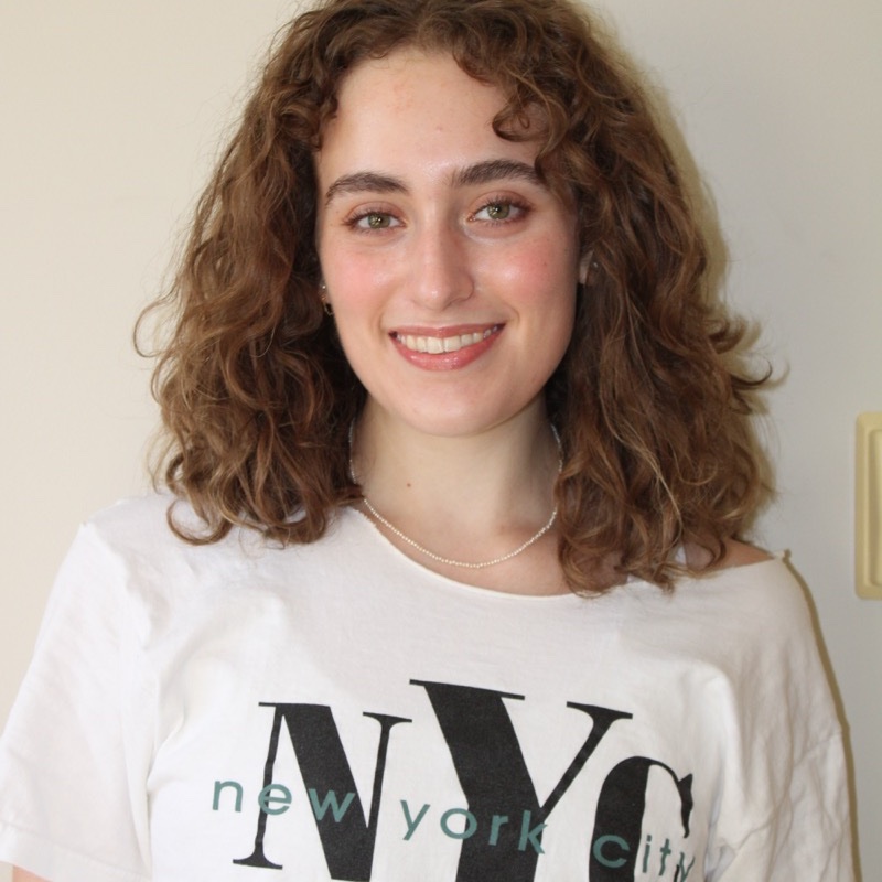 A person with curly, shoulder-length hair smiles while wearing an off-shoulder white shirt that reads “NYC” in large letters. A pearl necklace is visible around their neck. The lighting is natural, and the tone of the photo is casual. A light switch is partially visible on the right side.