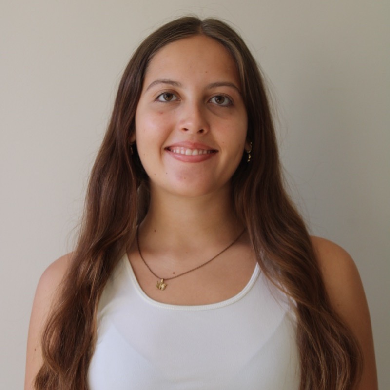 A person with long, softly waved hair stands against a plain background, smiling slightly. They wear a white tank top and a pendant necklace with a small charm. Their posture is relaxed and centered. The image is evenly lit with warm natural tones.