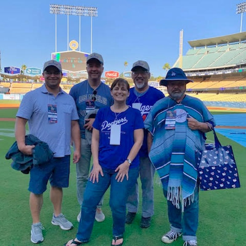  A group of five baseball fans poses for a picture on a baseball field. They are all wearing Dodgers-related clothing. The stadium stands, a scoreboard, and a bright blue sky are in the background.
