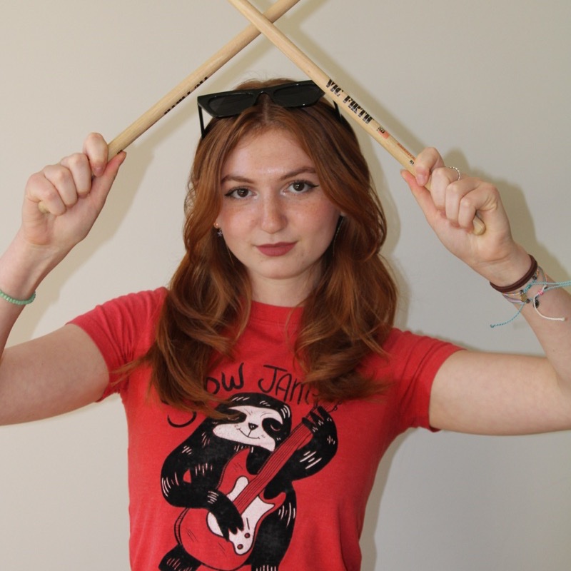 A person with long, wavy red hair holds two drumsticks crossed above their head. They wear a red t-shirt with a graphic of a sloth playing guitar and sunglasses resting on top of their head. Their expression is direct, and several bracelets are visible on their wrists. The background is a plain off-white wall.